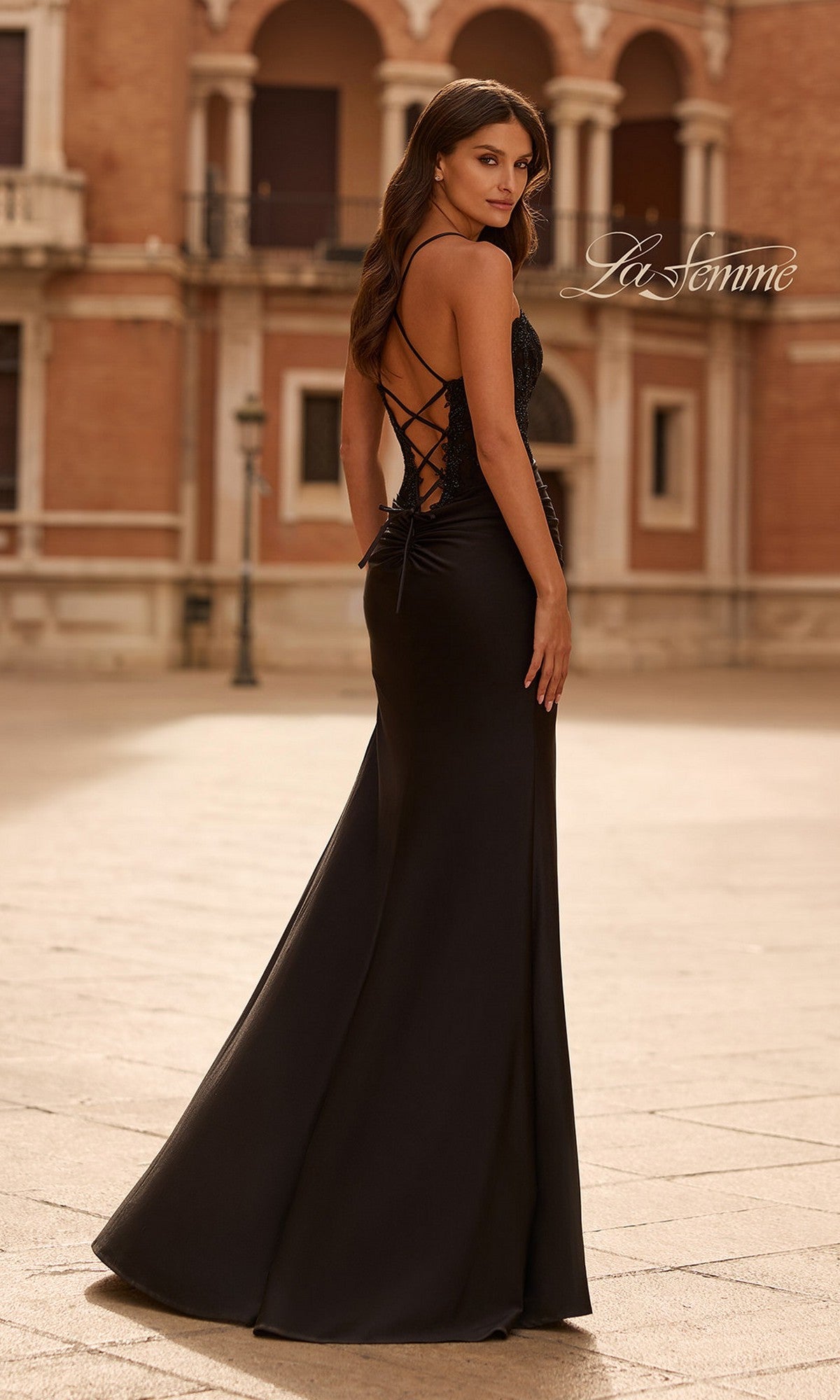 A woman stands outdoors on a stone plaza in front of a building, wearing the La Femme 33505 long prom dress—a black, backless evening gown with a plunging neckline and lace-up detailing.